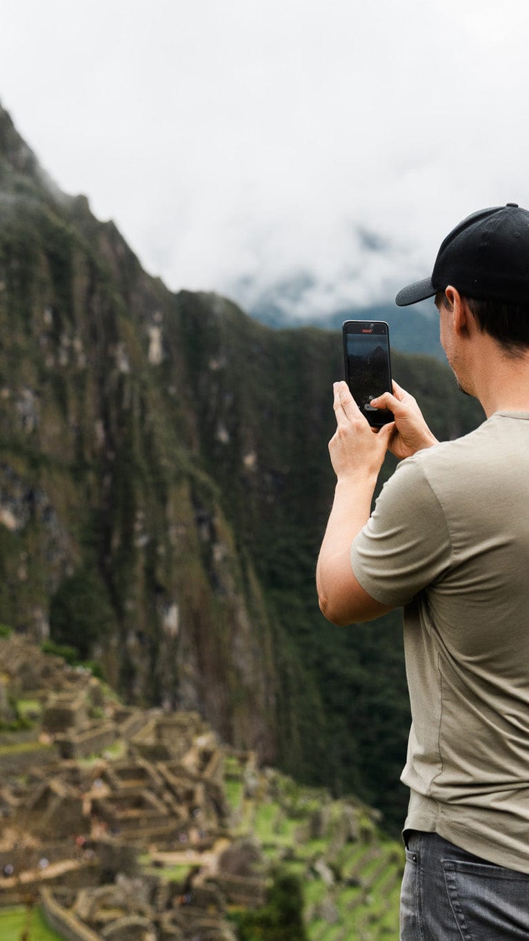 Young man in his mid-thirties wearing a green t-shirt and black baseball hat taking a phone video of the Machu Picchu – Travel video editing service.