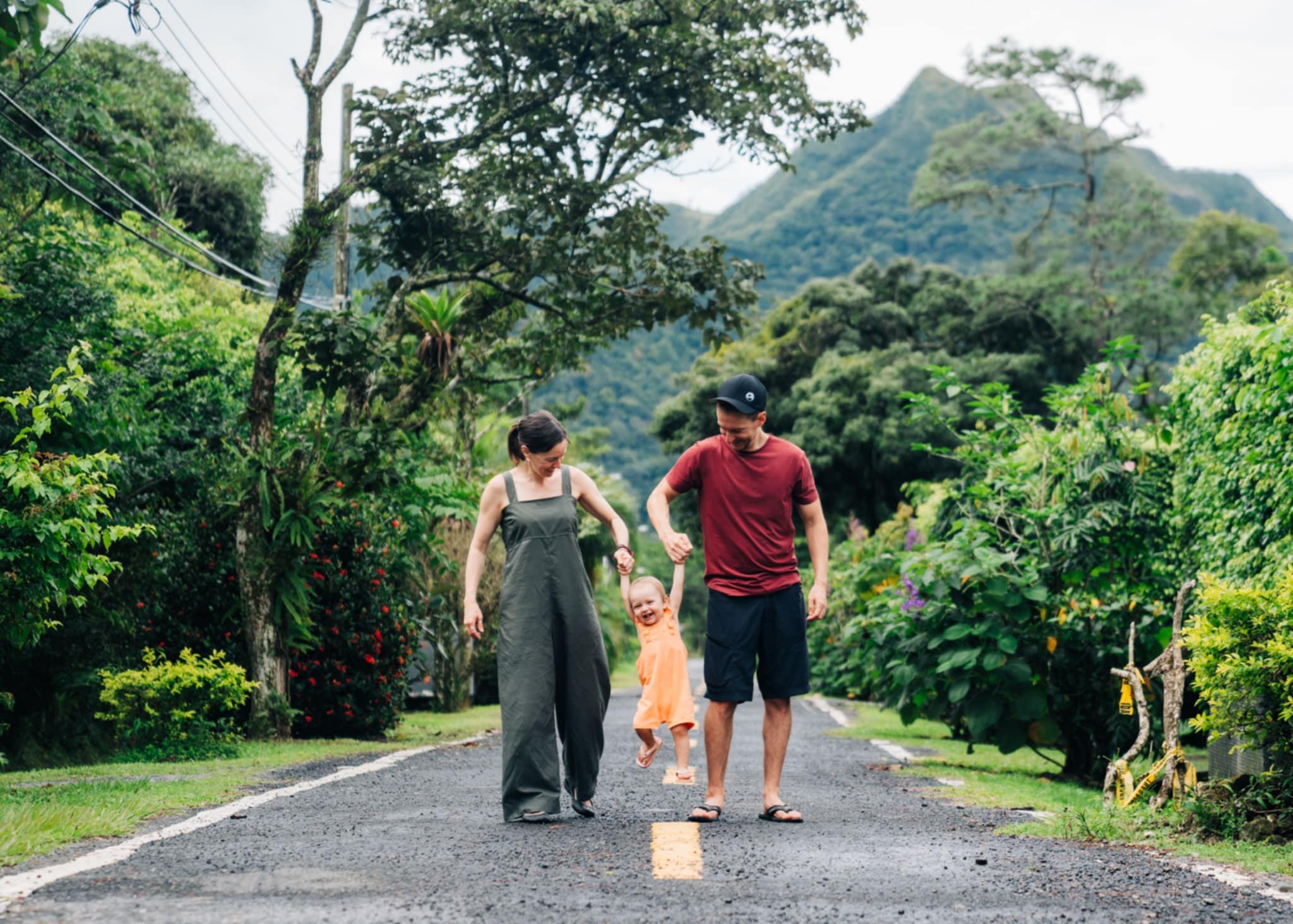 Young couple (Alex & MJ - On the GO) holding and lifting their baby daughter by the hands in the middle of a quiet street in Panama with many trees in the background – Family travel video editing service.