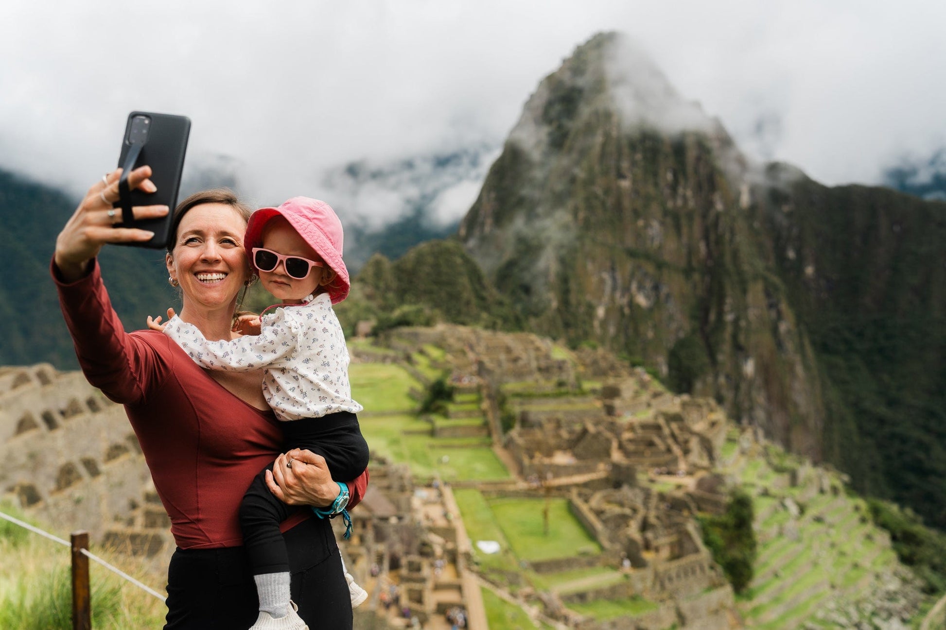 Woman holding her child and taking a selfie video with phone in front of the Machu Picchu.